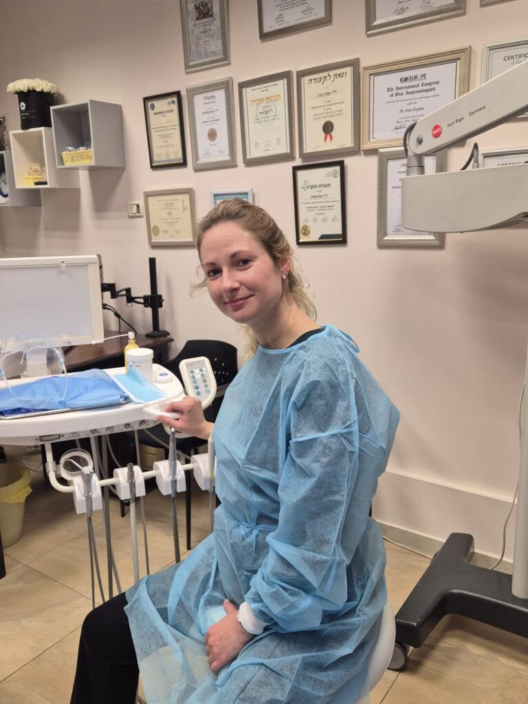 אישה לבושה חלוק רפואי כחול יושבת במרפאת שיניים, מוקפת בציוד רפואי ותעודות על הקיר.