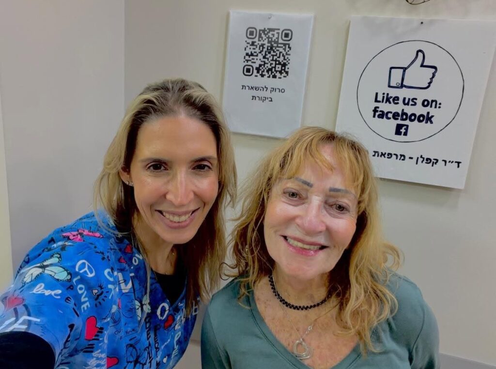 שתי נשים מחייכות בתוך מבנה ליד שלטים: באחד מהם מופיע קוד QR, ובאחר מפורסם דף פייסבוק של מרפאת שיניים מומלצת בפתח תקווה.