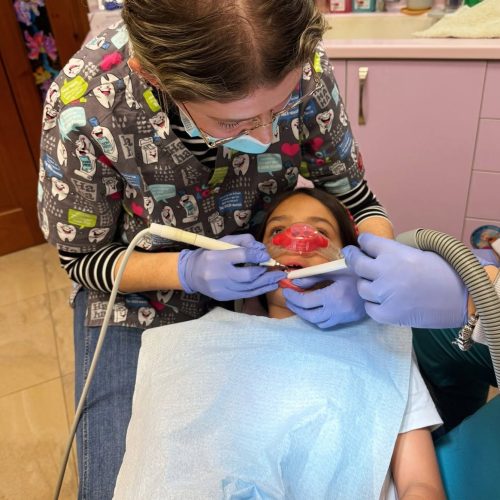 רופא שיניים, הלובש כפפות ומסכה, מבצע טיפול שיניים בילד, השוכב בכיסא רופא השיניים כשהוא חובש סינר ומסכת אף.